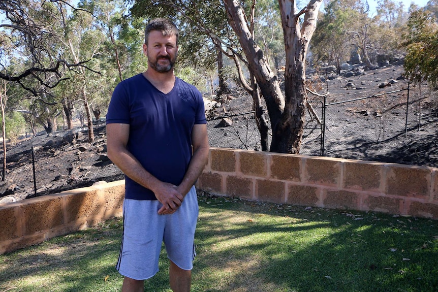 A man in the yard of his home, with bushfire blackened vegetation behind him.