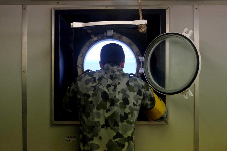 A man in a navy uniform stares out a porthole of a ship with his back to the camera