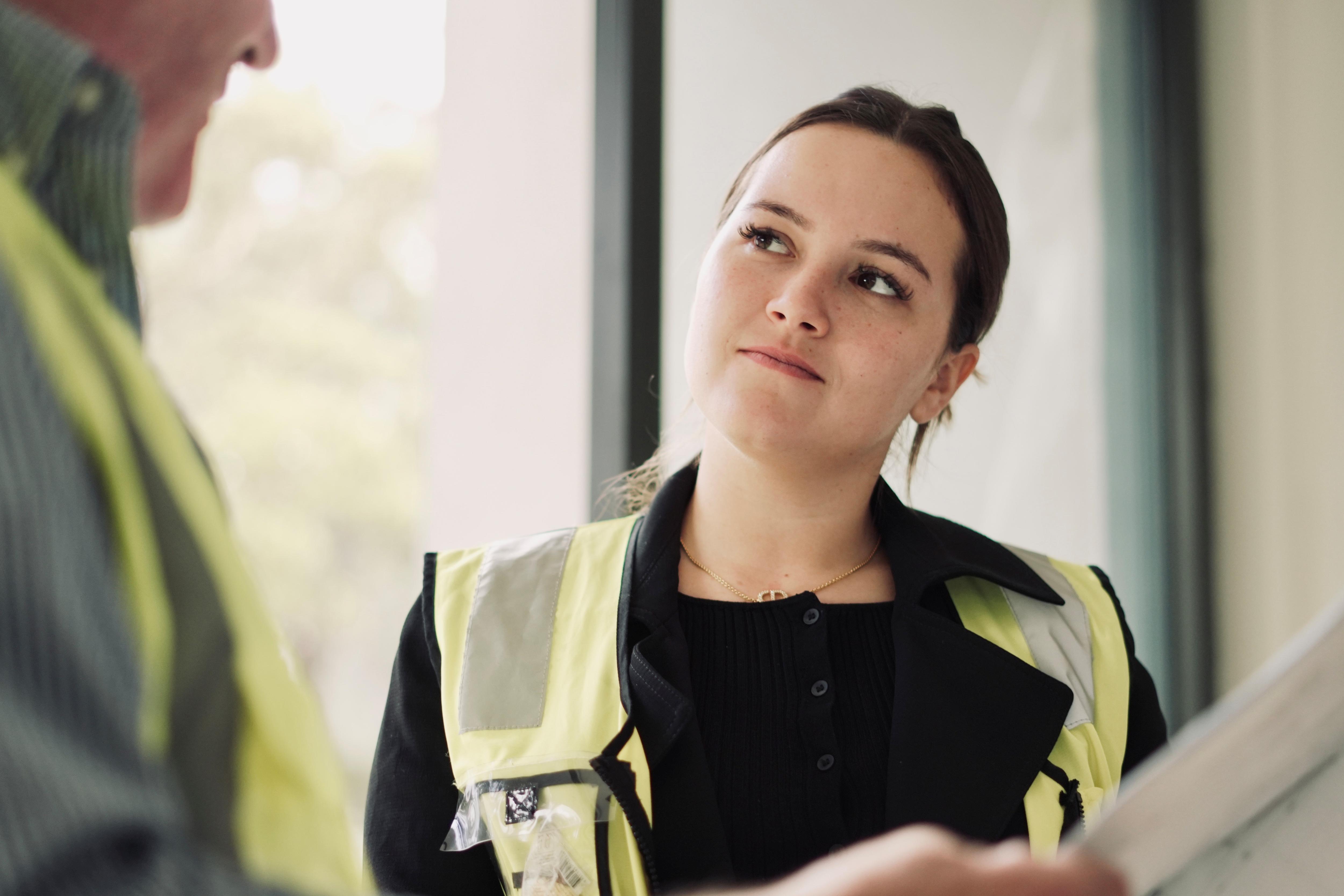 Slight smile, Paige in green hi viz vest listens intently to older man in green hi viz, shirt, face blurred and cut off. 