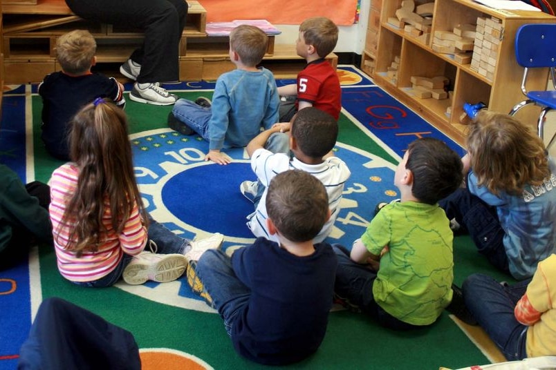 Young children sit on the floor of a classroom listening to their teacher