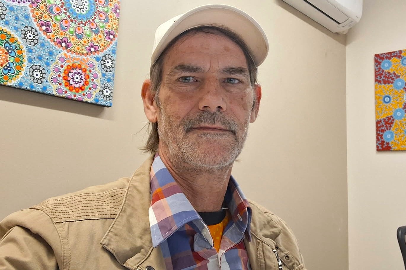 A man with gray hair and beard sitting in an office space, in a beige jacket and white cap looking directly at camera.