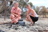 Two women crouched looking at camera, burnt cups and metal in foreground