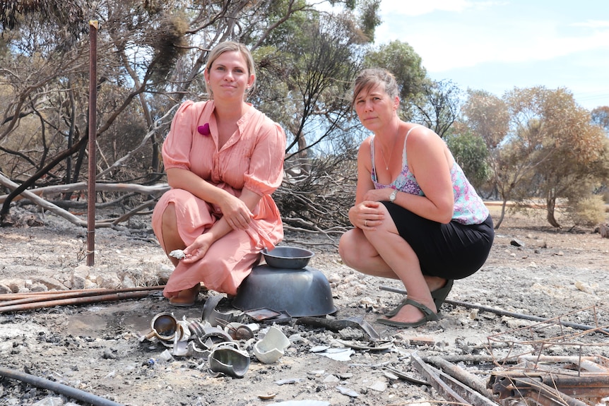 Two women crouched looking at camera, burnt cups and metal in foreground