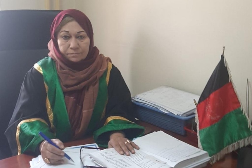 A woman sitting at a desk, wearing a headscarf and large coat, signs paperwork.