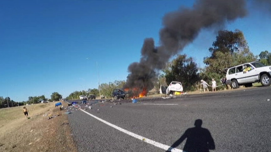 The wreckage of a dark coloured 4WD lies burning on the side of a road with a white SUV in a ditch nearby and debris scattered.