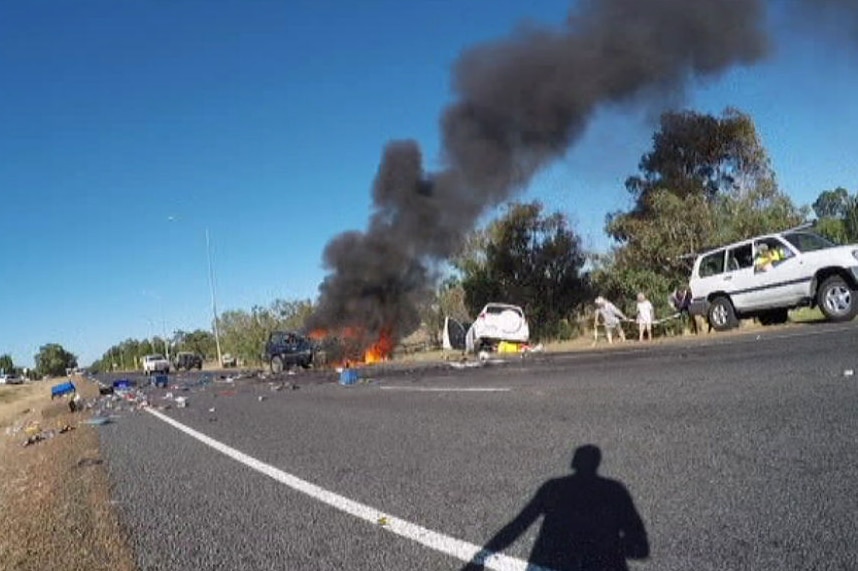 The wreckage of a dark coloured 4WD lies burning on the side of a road with a white SUV in a ditch nearby and debris scattered.