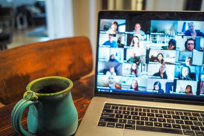 A green mug on a table in front of an open laptop with lots of faces on the screen.