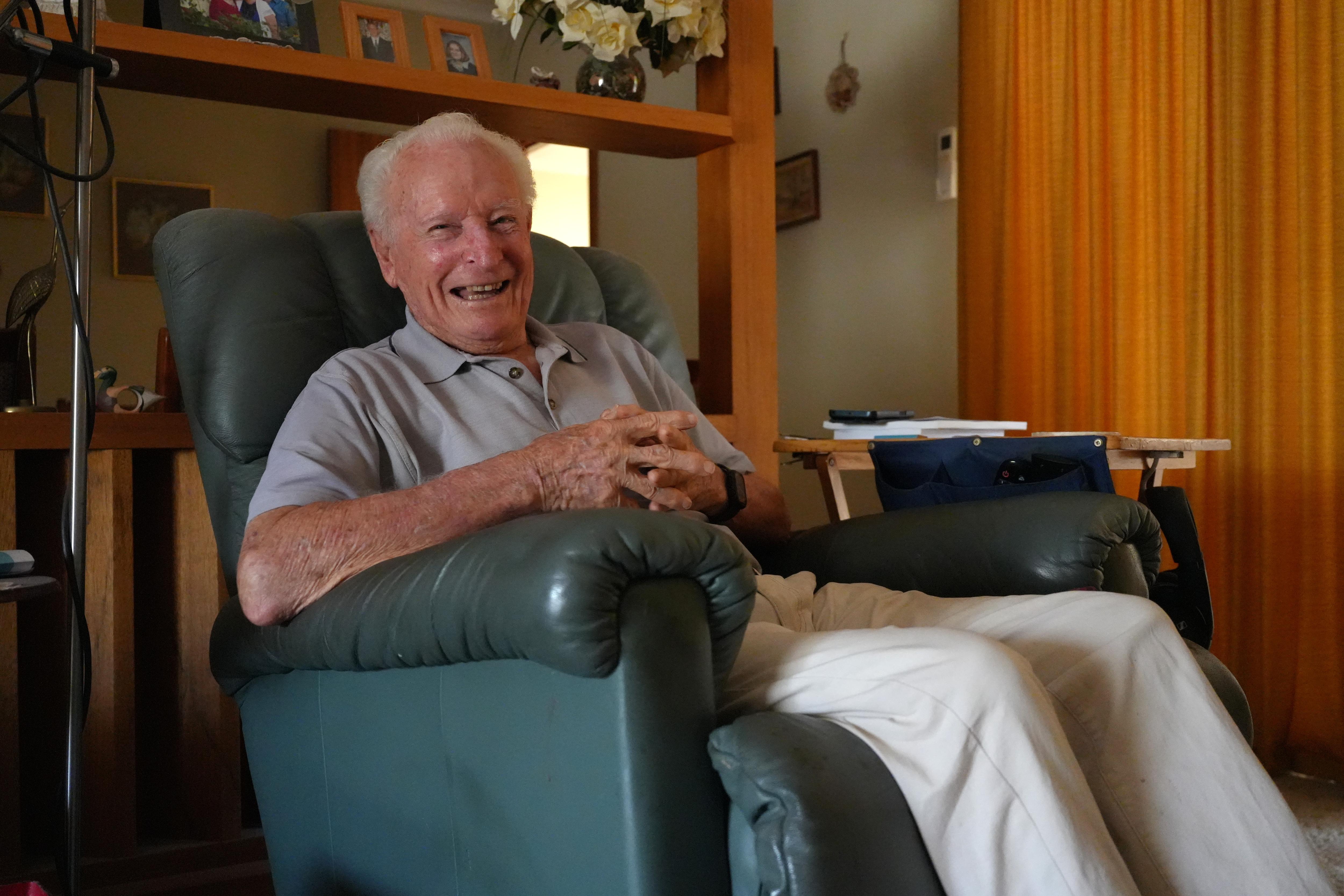 Clarence 'Clack' John está sentado em uma poltrona verde. Ele está sorrindo para a câmera e vestindo uma camisa pólo cinza e calça branca.