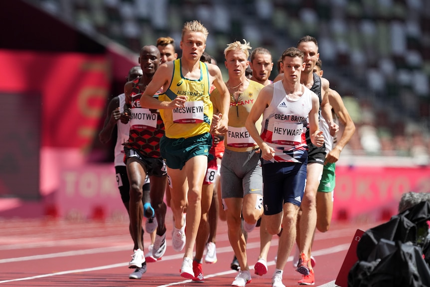 A blonde man wearing a yellow singlet runs on a track ahead of a group of men