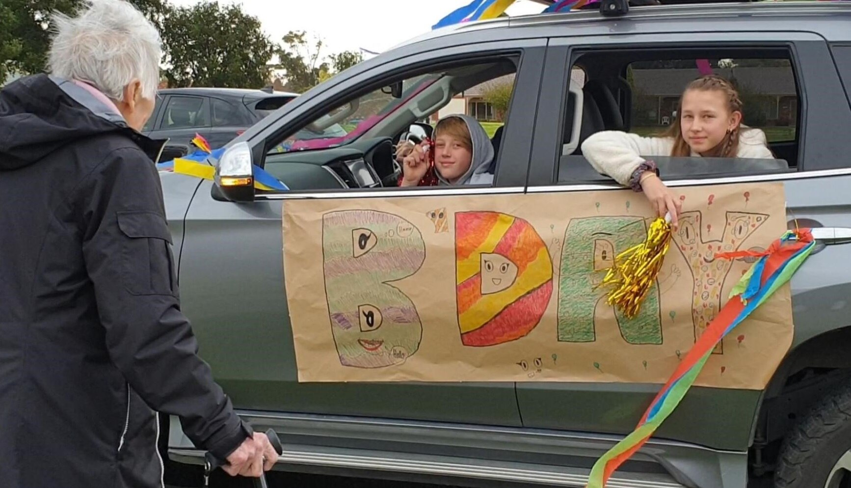 A grandmother looks on as her family drive past to celebrate her birthday.