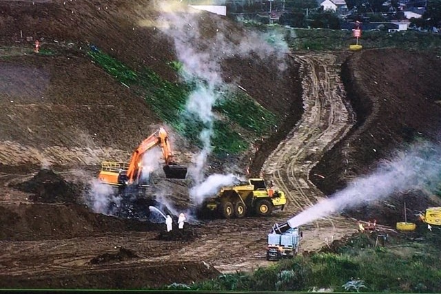 smoke rising from the ground at a tip, there are trucks working on the site nearby.