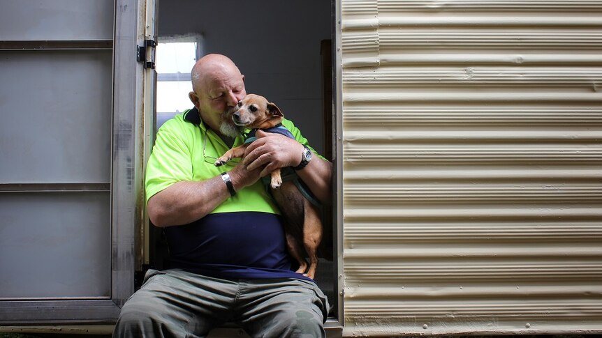 A man sits on the steps of a caravan, holding and kissing a small dog.