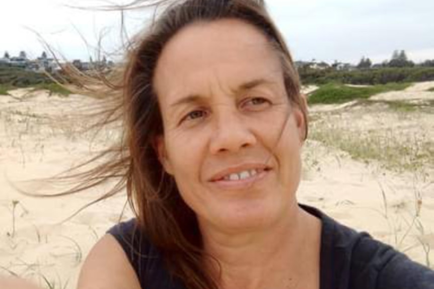 A smiling, middle-aged woman with dark hair takes a selfie on a beach.