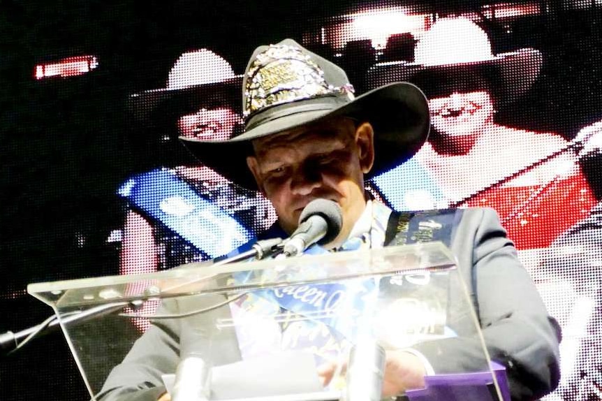 A man wearing a blingy cowboy hat stands on a stage, making a speech.