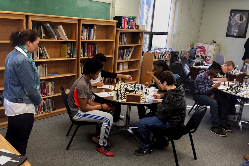 Two tables of children play chess in an informal setting while a woman supervises, with bookshelves in the back.