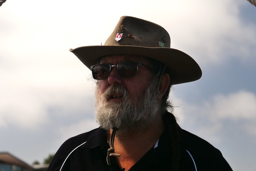 A man with a beard, hat and sunglasses in stark contrast to cloudy sky behind.