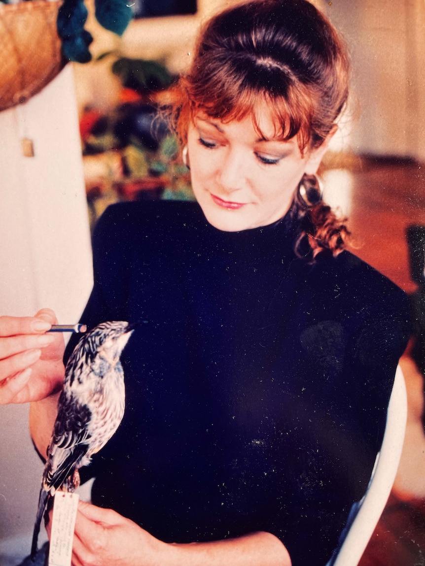 A woman in a black top looking down at a stuffed bird, holding a paint brush