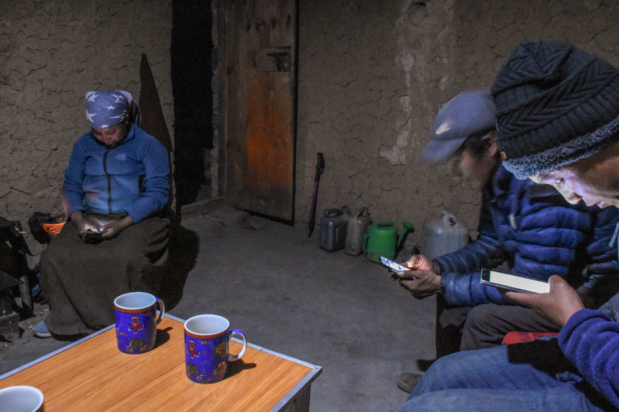 Two men and a woman stare at their smartphone screens inside a clay hut dimly lit