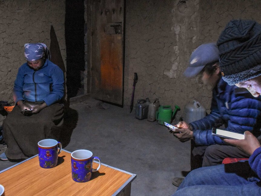 Two men and a woman stare at their smartphone screens inside a clay hut dimly lit