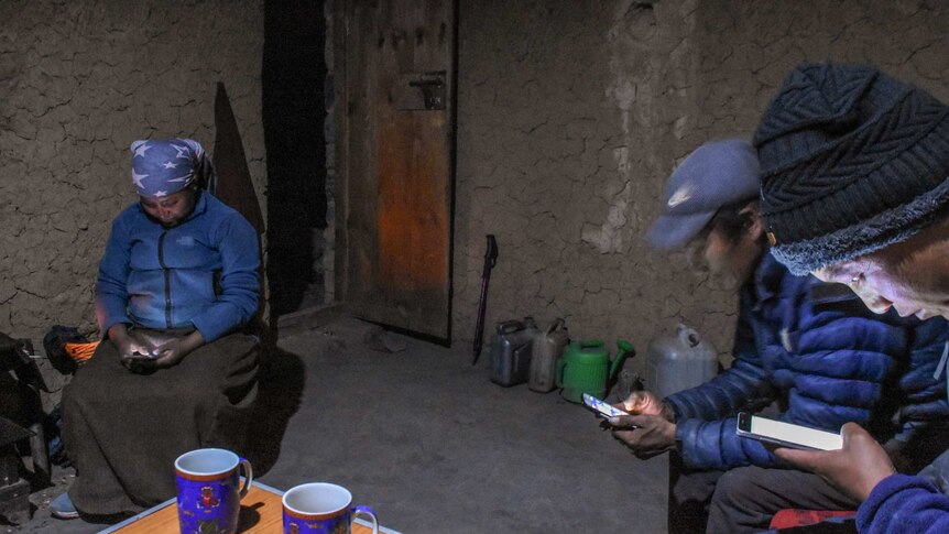 Two men and a woman stare at their smartphone screens inside a clay hut dimly lit