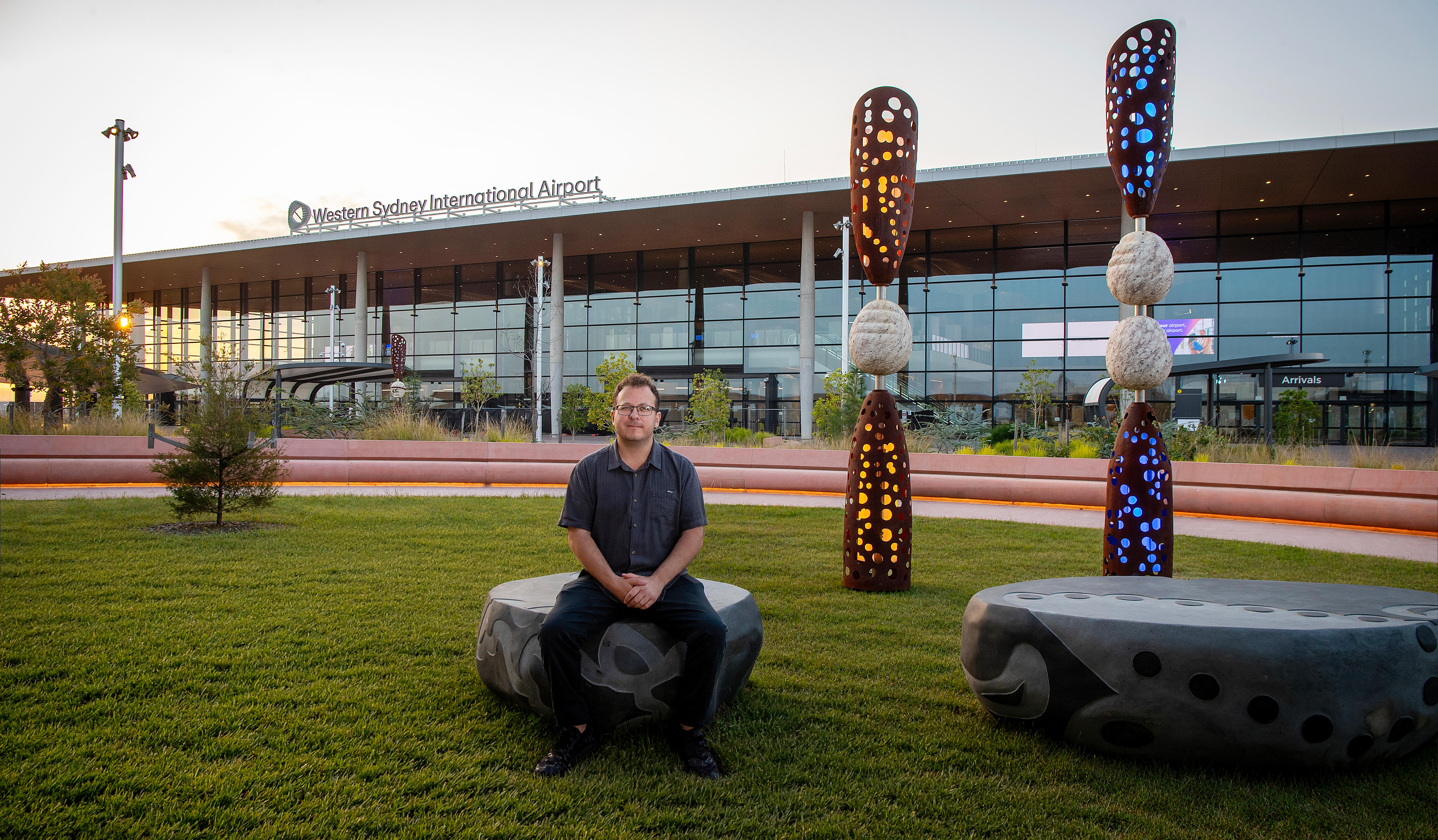 A man sitting outside an airport terminal called Western Sydney International, next to Indigenous sculture artwork, in the AM.