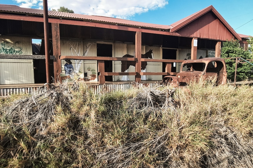A run-down outback pub with a rusted vehicle at rest among the weeds in front of it.