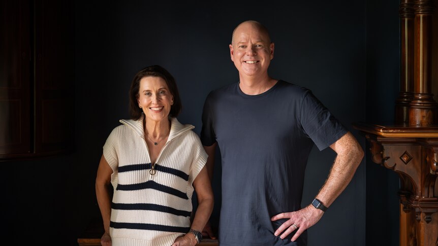 Virginia Trioli and Tom Gleeson, two middle-aged people, pose smiling together. Gleeson has a hand on his hip.