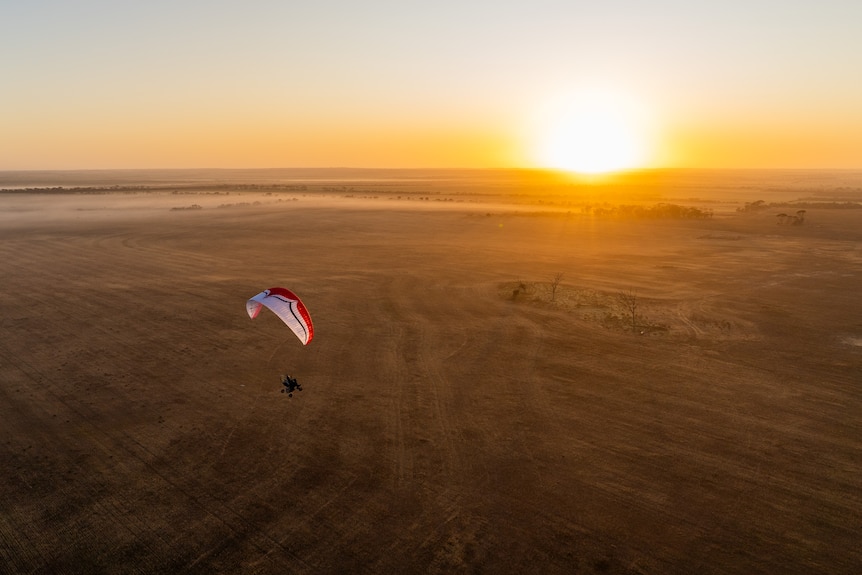 A man paramotoring over brown desert as the sun rises on the horizon.