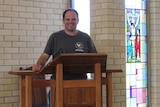Man stands at pew in a church with a stain glass window of Jesus on cross int he background.