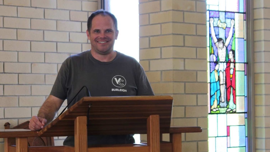 Man stands at pew in a church with a stain glass window of Jesus on cross int he background.