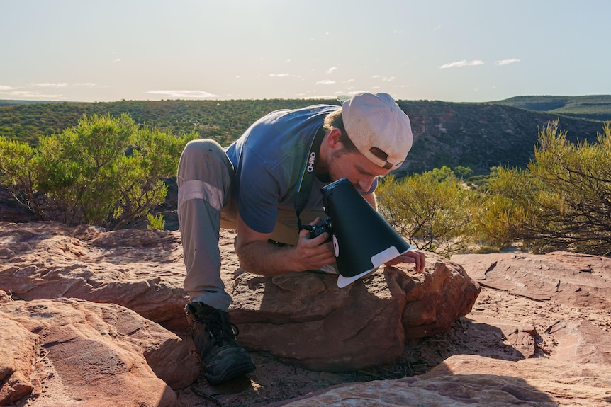 Flynn Prall kneels on a rock in the bush with his camera equipment.