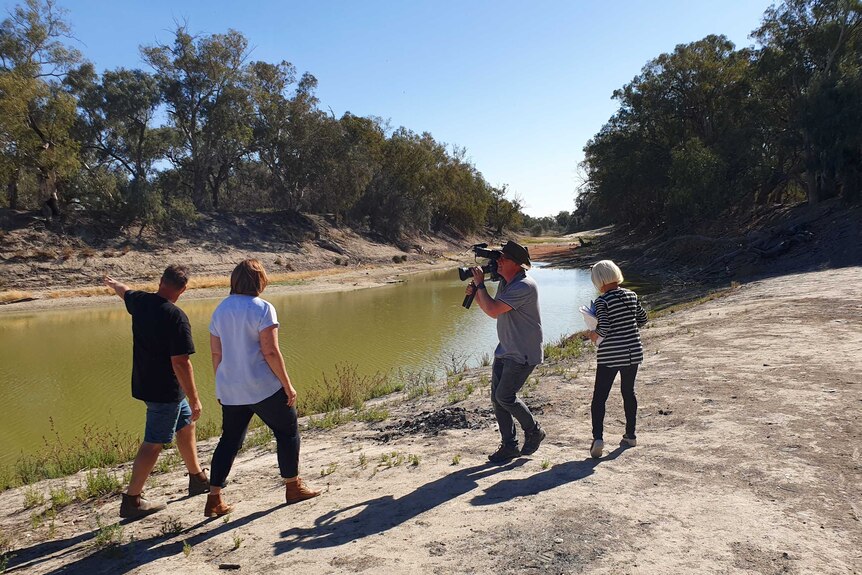 Cameraman holding camera, with producer standing behind, filming Millar and a man walking along dry river bank with low water.