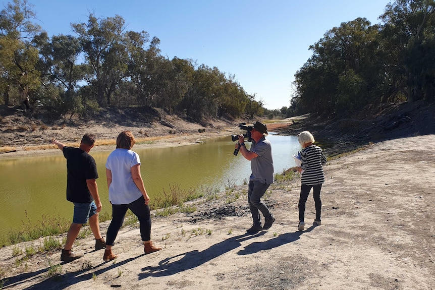 Cameraman holding camera, with producer standing behind, filming Millar and a man walking along dry river bank with low water.