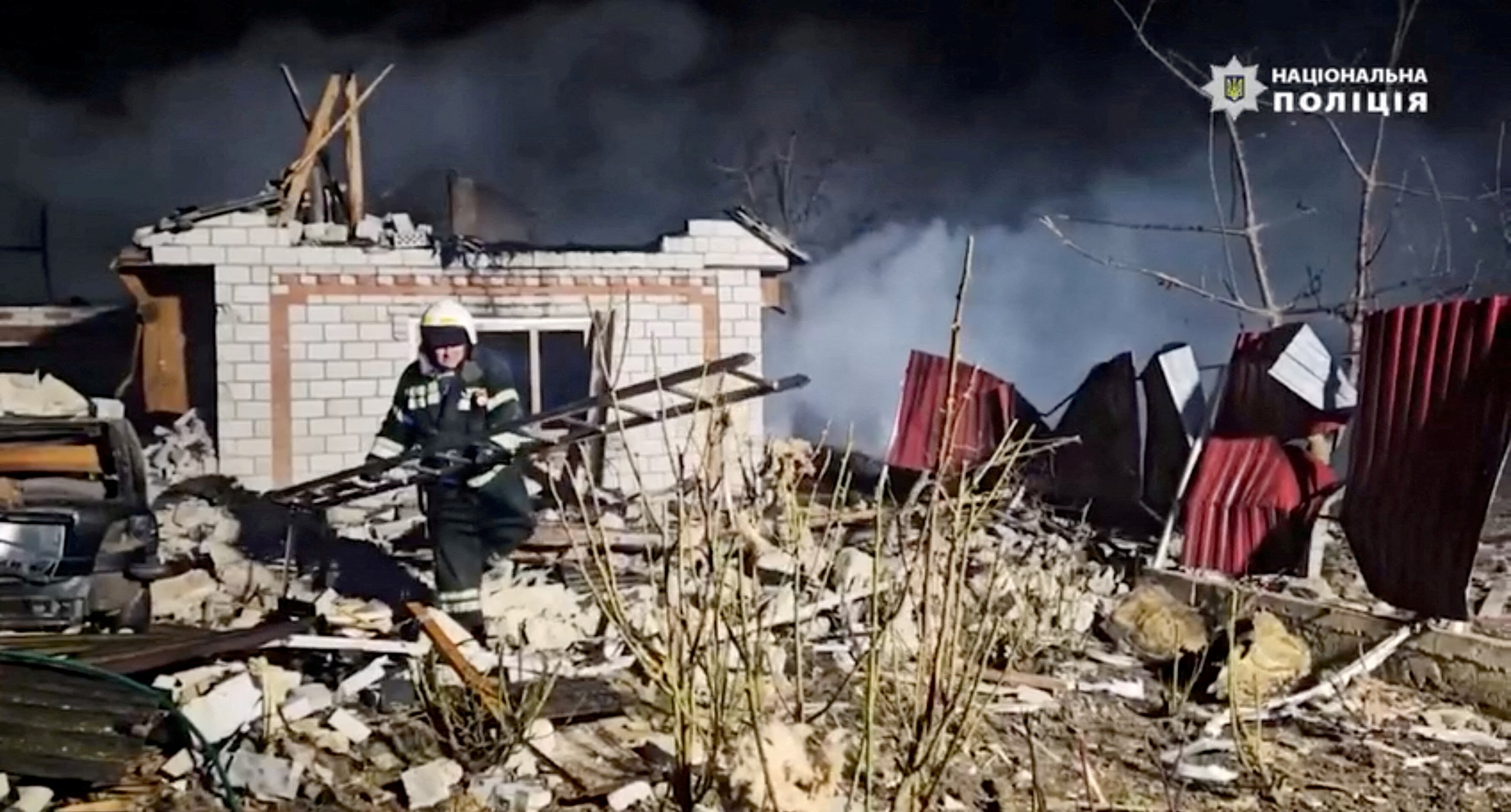 An emergency service worker steps over rubble of former home carrying a ladder with smoke billowing in the background