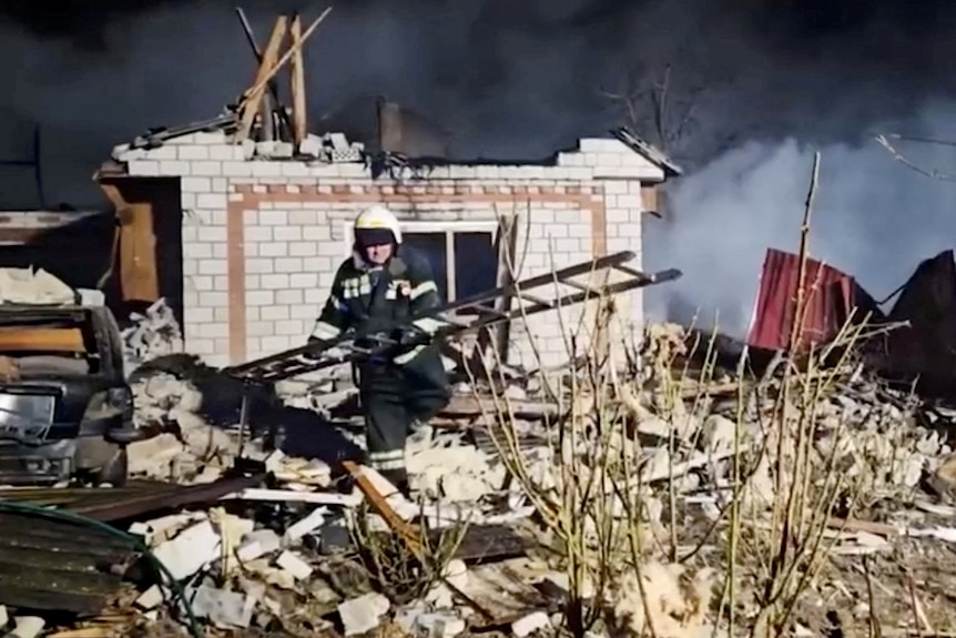 Pracownik służb ratunkowych przechodzi przez ruiny dawnego domu, niosąc drabinę, a w tle kłębi się dym