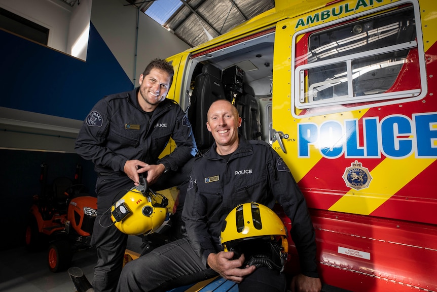 Two police officers holding helmets sit beside a rescue helicopter