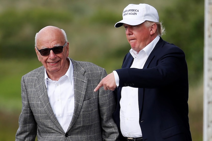 Rupert Murdoch and Donald trump walk side by side. Murdoch is smiling, Trump points at Murdoch while wearing a white MAGA hat.