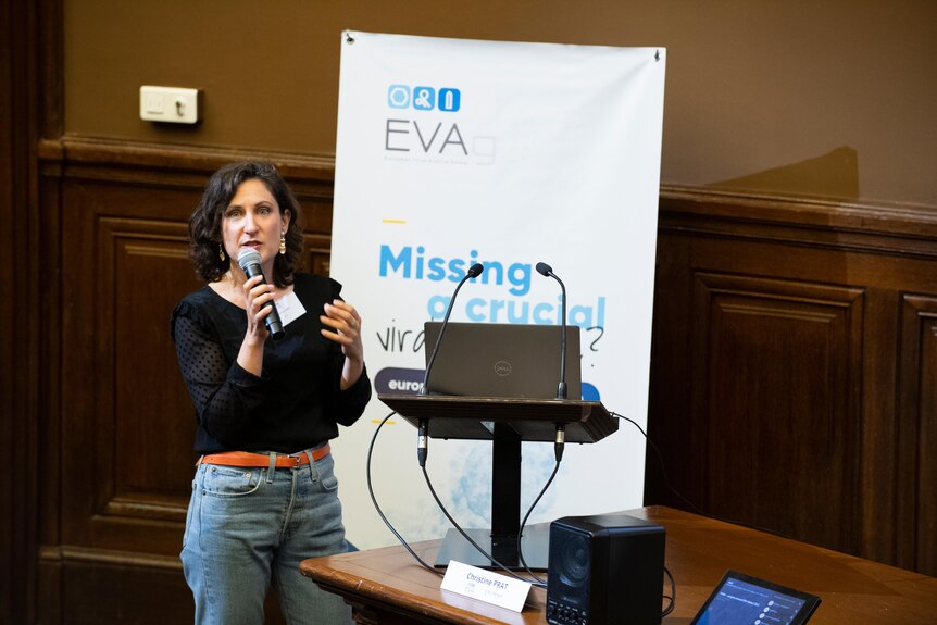 A woman with wavy dark hair, black top and jeans gives a speech in front of a laptop, podium and banner inside a room