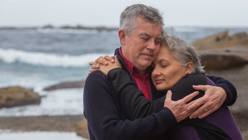 A man and woman embrace, with eyes closed, on a rocky beach.