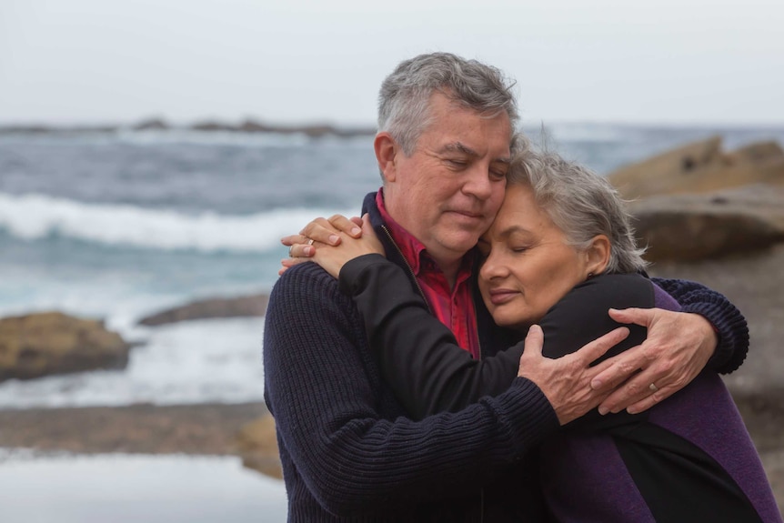 A man and woman embrace, with eyes closed, on a rocky beach.