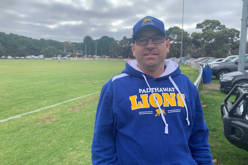 A man in a matching blue and yellow jumper and cap leans against a fence around a football oval.