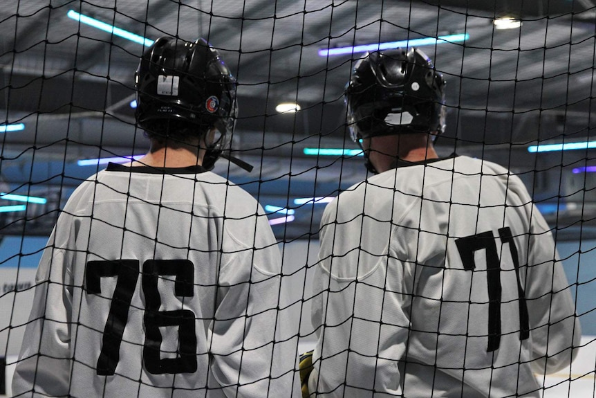 Two teammates stand at the side of the rink watching the game.