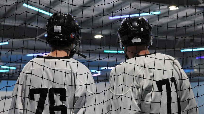 Two teammates stand at the side of the rink watching the game.