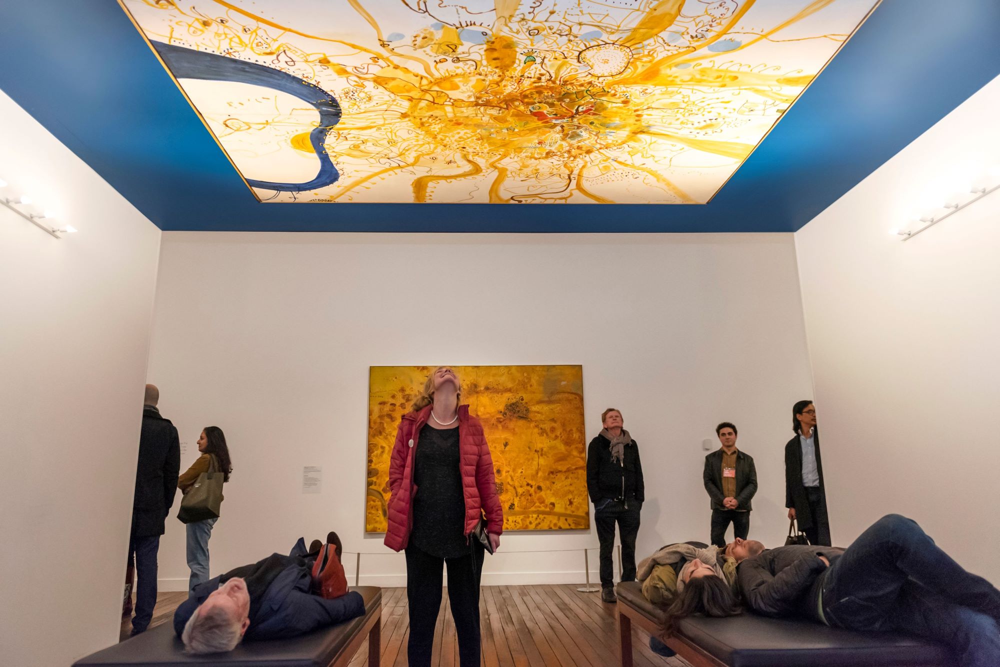 In a gallery a group of people stand or lie down all looking up at an abstract landscape painting in white, yellow and blue