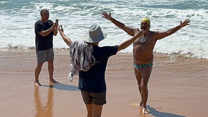 a man in swimming costume stretches hands outwards on a beach