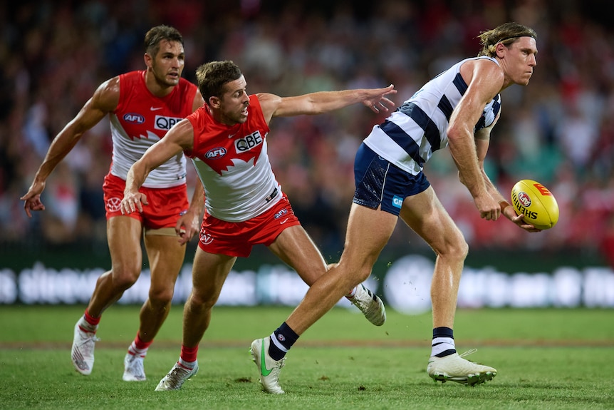 A tall Geelong ruckman goes for a handball in the middle of the ground as two Sydney defenders scramble to stop him.