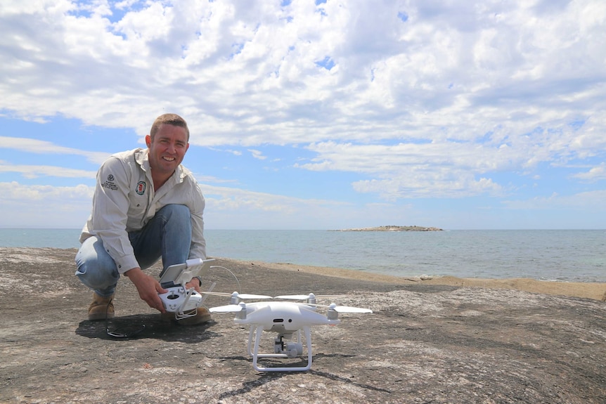 man looking at camera holding drone controls, drone on ground with ocean and island in background