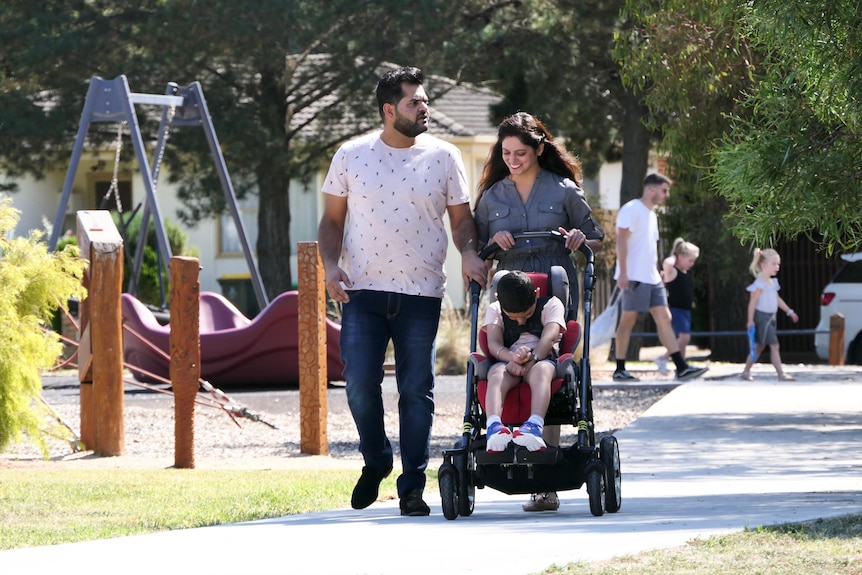 Priyanka is pushing a pram and smilng down at her son Kayaan, walking next to Varun on a footpath near a playground.