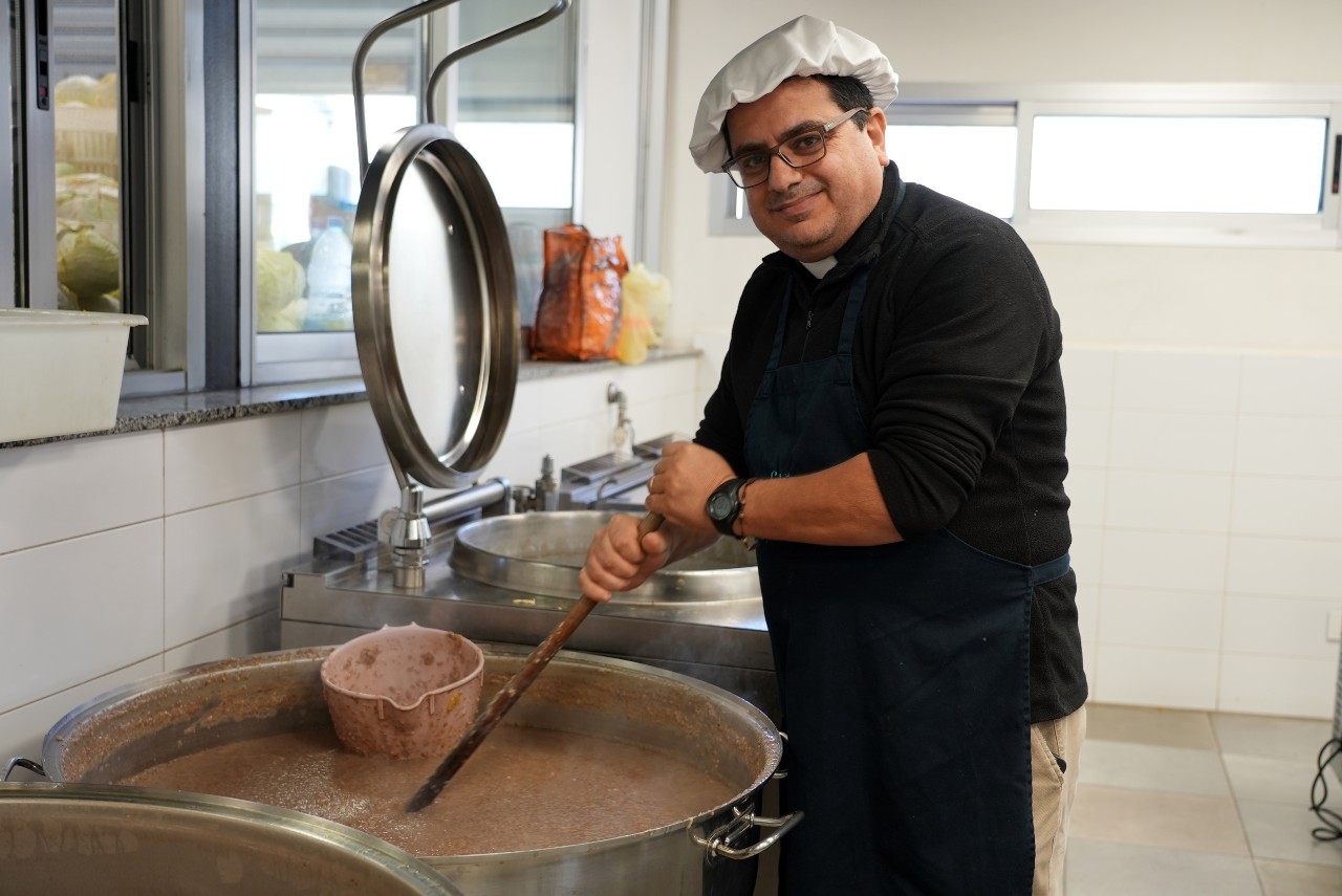 A man smiling at the camera while stirring a large pot of food.