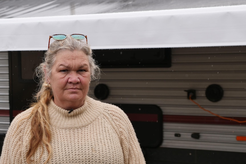 A woman in a fleece jumper stands in front of a caravan in the rain.